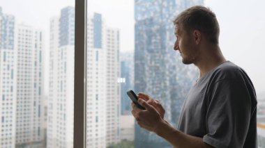 Beyaz adam gökdelen dairesinden kentsel manzaranın tadını çıkarırken akıllı telefonuna göz atıyor. 