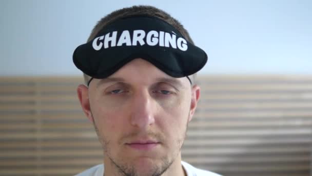 Exhausted and tired man puts on a sleeping mask with charging written on it and goes to bed — Stock Video