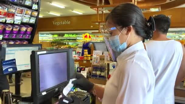 Mujer cajero asiático en máscara protectora mirando a un cliente en el supermercado. Compras esenciales durante el brote de coronavirus. Bali-junio-2021 — Vídeos de Stock