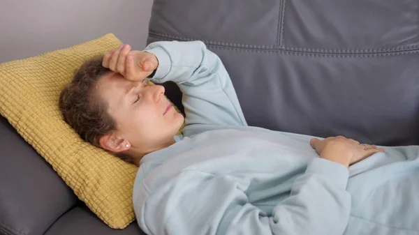 Egy fiatal nő a kanapén fogdossa a fejét, hogy magas-e a láza. Fejfájás és láz fogalma — Stock Fotó