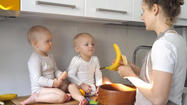 Jovem Multitasking mãe alimenta banana seus bebês gêmeos enquanto faz um jantar — Fotografia de Stock