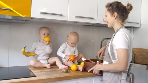 A vida real de uma mãe solteira. Bebês brincando com frutas sentadas na mesa enquanto ela mistura o molho — Fotografia de Stock