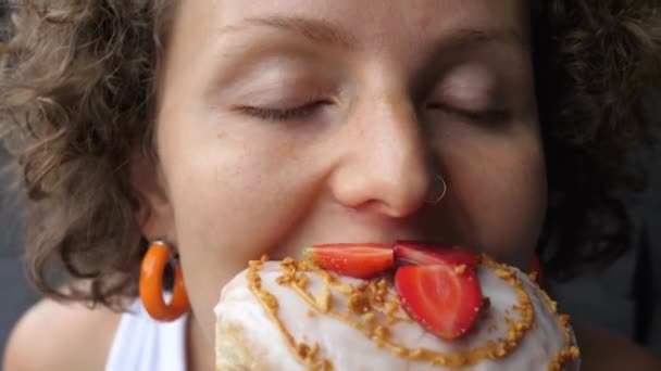 Närbild av lockigt kaukasiska kvinna med ätstörningar biter en munk toppad med jordgubbar aggressivt och tuggar den snabbt — Stockvideo