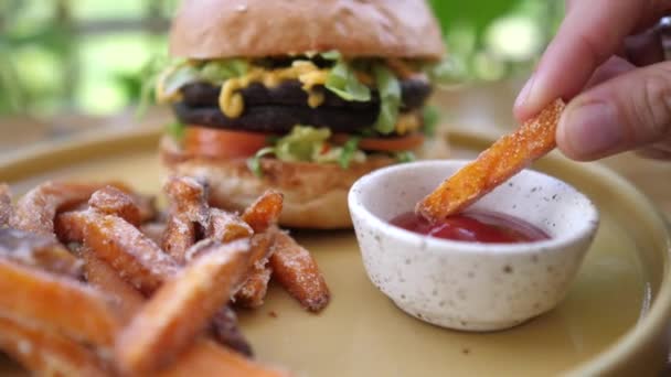 Dipping sweet potato in ketchup. Plant based burger in the background. Healthy vegan lunch in american style — Stock Video