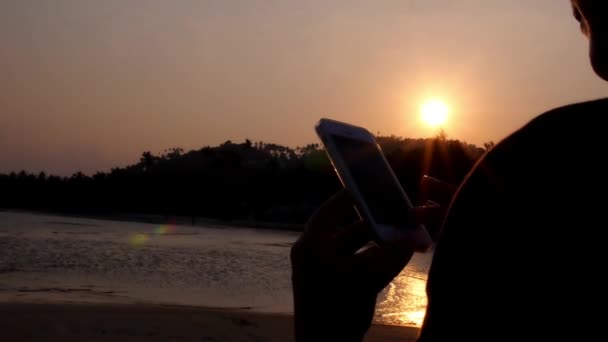 Mão com telefone celular contra o pôr do sol na praia . — Vídeo de Stock