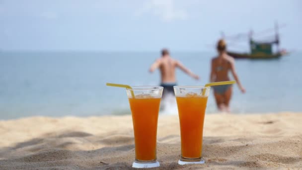 Dois copos de suco de laranja na praia e casal de natação — Vídeo de Stock