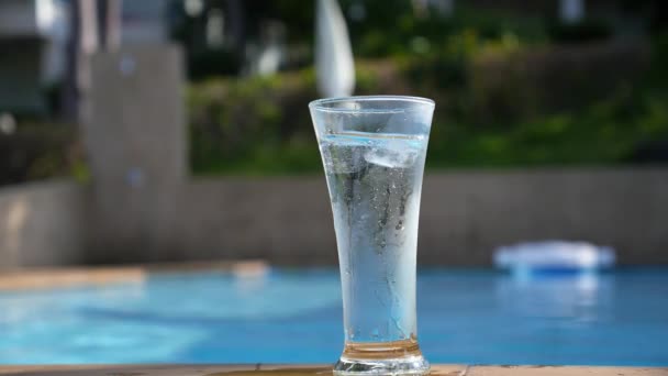 Bicchiere d'acqua con cubetti di ghiaccio sul lato della piscina blu — Video Stock