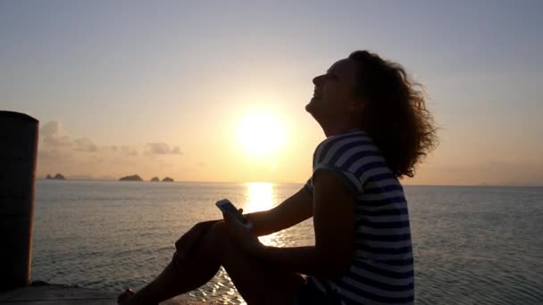Mulher usando telefone celular e rindo no pôr do sol do mar — Vídeo de Stock