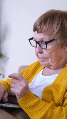 Kanepede oturan ve evinde dizüstü bilgisayar kullanan yaşlı bir kadın, ekranda konsantre olurken interneti gezmek için parmağıyla dokunuyor.