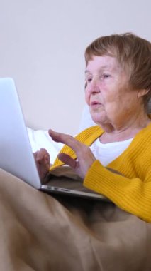 Yaşlı kadın, ailesiyle video görüşmesi yapmak için dizüstü bilgisayarını kullanarak yatakta uzanıyor. Modern teknoloji sayesinde evindeki konforunu koruyarak gülümsüyor ve konuşuyor.