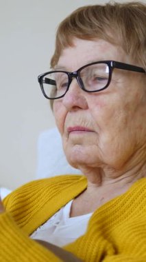 Gözlüklü yaşlı kadın akıllı telefonunu dikkatli bir şekilde kullanmayı öğreniyor, rahat bir koltukta otururken ekrandan süzülüyor, modern dijital teknolojide ustalaşmaya odaklanıyor.