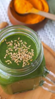 Keten tohumlarıyla süslenmiş ve sağlıklı, antioksidan zengini bir kahvaltı için parlak sarı zerdeçal tozunun yanında sunulan mason kavanozundaki lezzetli yeşil smoothie.