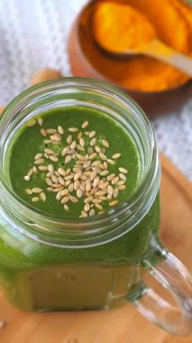 Sağlıklı yeşil smoothie, cam kavanozda susam tohumları ile kaplı, bir kase zerdeçal tozuyla ahşap bir tahtanın üzerinde dönüyor, besleyici ve vegan dostu bir kahvaltı sergiliyor