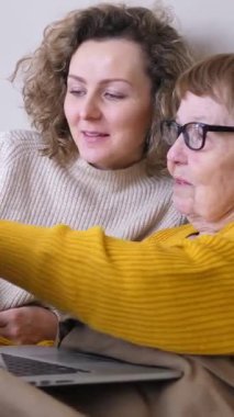 Genç bir kadın, yaşlı kadın internette gezinmeyi ve ailesiyle iletişimde kalmayı öğrenirken dizüstü bilgisayarını nasıl kullanacağını gösterip ekranı işaret ediyor.