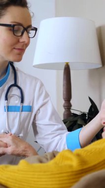 Genç bir bayan doktor steteskopla yaşlı bir kadını ev ziyareti sırasında rahatlatıyor, tıbbi danışmanlık ve tedavi sırasında ona destek olmak ve şefkat göstermek için elini tutuyor.