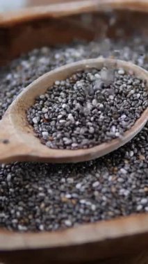 Besleyici chia tohumlarının yakın çekim görüntüsü yavaşça bir kaseye konmuş ahşap bir kaşığa düşüyor, dengeli bir diyet için sağlıklı beslenme ve süper yiyecek kavramını vurguluyor.
