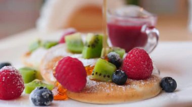 Lezzetli bir kahvaltı için ahududu, yaban mersini, kivi dilimleri ve pudra şekerle süslenmiş taze yapılmış krep yığınının üzerine dökülen tatlı bal veya akçaağaç şurubu.