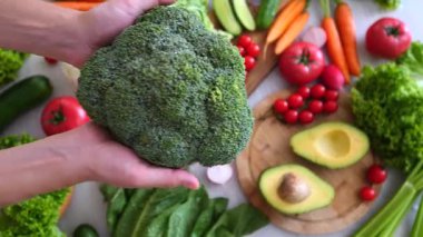 Renkli sebzelerle dolu bir masanın üzerinde taze organik brokoli tutan kadın elleri sağlıklı bir yaşam tarzını, vejetaryen diyetini ve doğal malzemelerle temiz yemek yemeyi destekliyor.