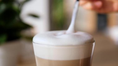 Bir bardak lezzetli cappuccino ya da latte macchiato 'dan kaşık yavaşça kremalı beyaz süt köpüğünü kaldırarak yumuşak ve odak dışı bir arka plana karşı güzel ve narin bir şekil oluşturur.