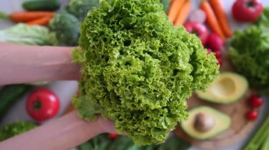 Kadın eli, taze yeşil marulu rengarenk organik sebzelerle dolu bir mutfak masasına koyar. Dengeli bir diyet için sağlıklı bir beslenme tarzı ve vejetaryen yemeği gösterir.