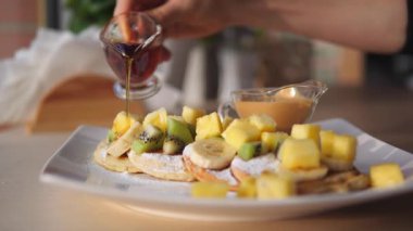 Cam sürahide tatlı akçaağaç şurubu dökülen kişi taze yapılmış krepler, lezzetli bir şekilde ananas parçaları, dilimlenmiş kivi, muz ve pudra şekeri ile süslenmiş