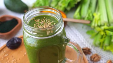 Susam tohumlarıyla kaplı cam mason kavanozunda taze yapılmış yeşil smoothie. Etrafı kereviz, avokado, hurma, ceviz ve detoks diyeti için spirulina tozu gibi sağlıklı maddelerle çevrili.