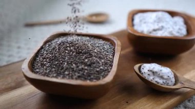 Sağlıklı chia tohumlarının yavaş çekimde, yakınlarda bir kaşıkta kremalı puding ile kırsal ahşap bir kaseye dökülmesi, besleyici bir süper yiyecek kahvaltı malzemesi sergiliyor.