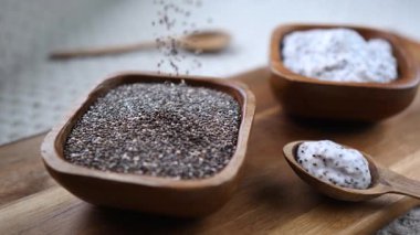Besleyici chia tohumları kırsal bir ahşap kaseye dökülüyor kaşıkta lezzetli chia pudinglerinden servis edilen tabağın yanında, sağlıklı bir süper yemek hazırlığı sergiliyor.