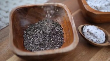 Çiğ chia tohumları, arka planda bir kaşık dolusu hazır chia pudingi bulunan, sade bir ahşap kaseye dökülüyor ve sağlıklı bir süper yiyecek içeriğini vurguluyor.