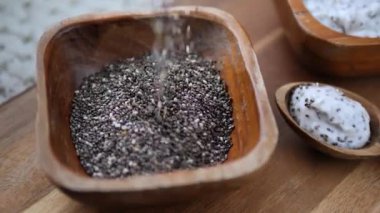 Besleyici çiğ chia çekirdekleri yavaşça kırsal ahşap bir kaseye dökülüyor, kaşığın üzerinde Chia pudingi bitmiş, sağlıklı beslenme ve süper yiyecek hazırlığı kavramını vurguluyor.