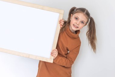 Mutlu küçük kız gülümsüyor ve elinde boş bir çizim tahtası var. Beyaz sırt çantasındaki test için kopyalama alanı.