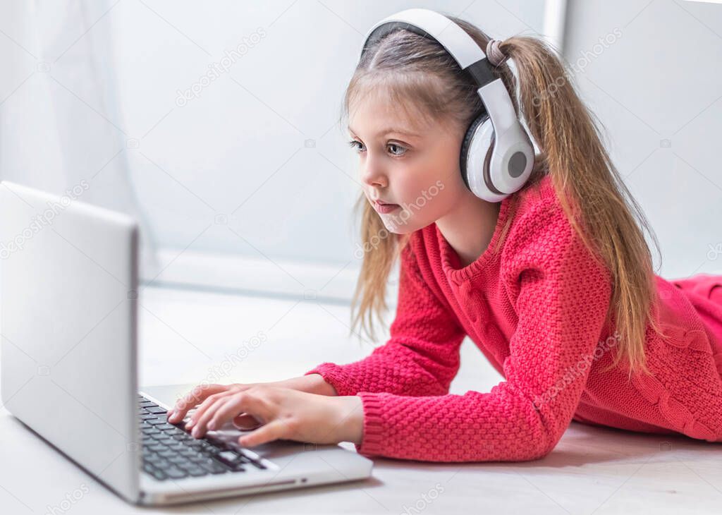 Cute little girl in headphones seriously using laptop at home for online education, home studying and online communication. Kids distance learning. Social distance. Stay at home.