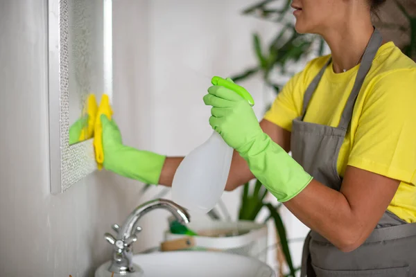 Cleaning the bathroom. The girl rubs the sink and mirror. Hotel service. Professional cleaning of apartments and premises. Yellow and green uniforms. Rubber gloves, apron and cleaning supplies