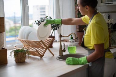 Mutfağın genel temizliği. Önlüklü ve lastik eldivenli bir kadın lavaboyu yıkıyor. Profesyonel ev işi. Bulaşıkları bezle siler