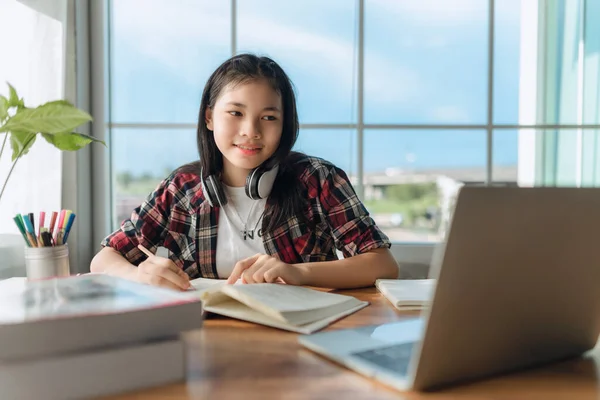 Asian teen girl wearing headphones learning language online, using laptop, looking at screen, doing school tasks at home, writing notes, listening to lecture or music, distance education