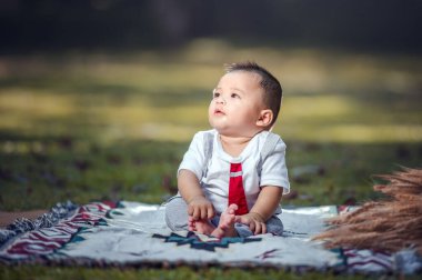 A little boy is sitting on a cloth spread on the grass in the park. 6 month old boy.