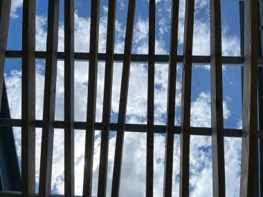 Parkta parlak mavi gökyüzü ve beyaz kabarık bulutları olan gri ahşap bir pergola çatısı..