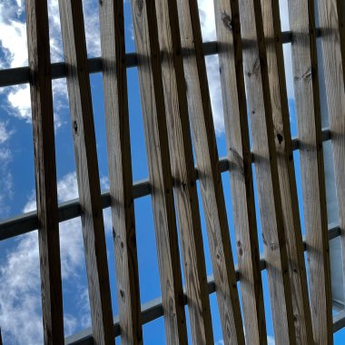 Parkta parlak mavi gökyüzü ve beyaz kabarık bulutları olan gri ahşap bir pergola çatısı..