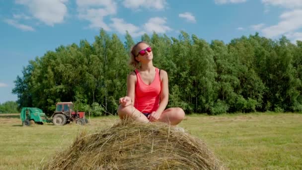 Beautiful girl on a sunny day in red T-shirt and glasses sits on haystack smiles — Stock Video