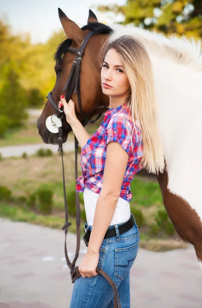 Menina bonita vestindo camiseta e jeans montando um cavalo no dia ensolarado de verão. Cabelo longo loiro com e um sorriso feliz. Pele perfeita e maquiagem. Fecha. Tiro exterior . — Fotografia de Stock