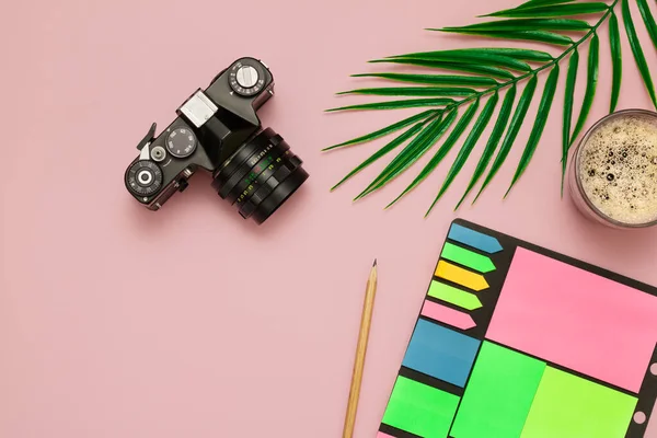 A camera, a notebook with a pencil, a cup of coffee and a leaf of a palm tree lie on a pink background with an empty place for the text. Concept - vacation or work, business, top view, layout.