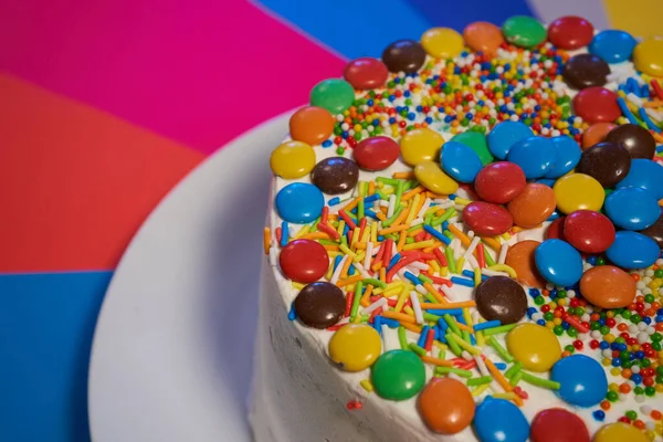 Beyaz bir tabakta servis edilen çok renkli şekerlemelerle kaplı kek ve krema. gökkuşağı arka planı. doğum günü ya da tatil