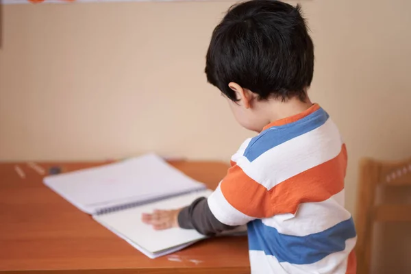 Sırtı kameraya dönük çizgili tişörtlü çocuk çalışma defterinde hareketler yapıyor.