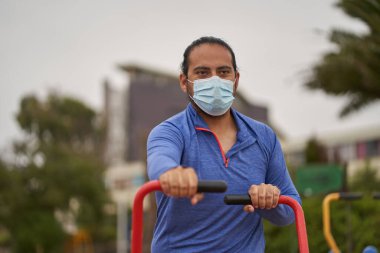 Mavi tişörtlü ve çene bantlı adam halka açık alanda makineler üzerinde kol egzersizi yapıyor. Yeni normalde maske kullanımı. spor oturumu.