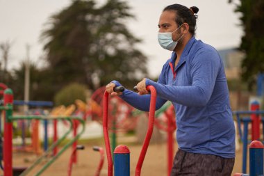 Yüz maskesi takan kıllı adam halka açık meydanda makineler üzerinde eliptik egzersizler yapıyor. yeni normal. spor oturumu