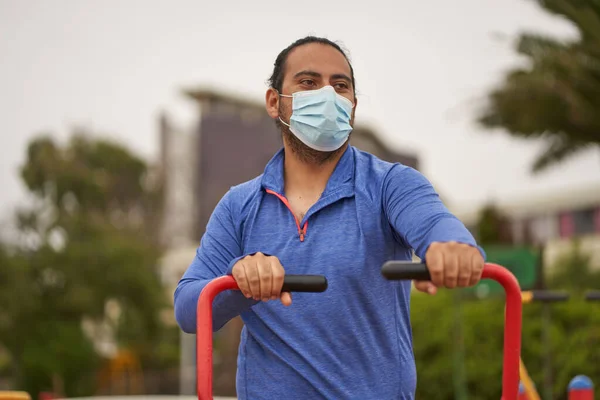 man performs arm exercises on machinery in public square. use of mask in new normal. sports session.