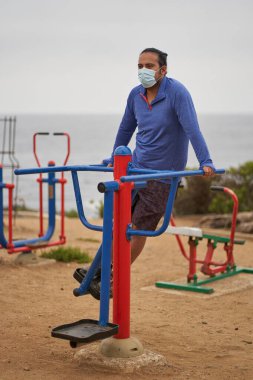 Yeni normal. Maskeli ya da çene bantlı bir adam parkta bel egzersizi yapıyor. Egzersiz makineleriyle kare. Spor oturumu