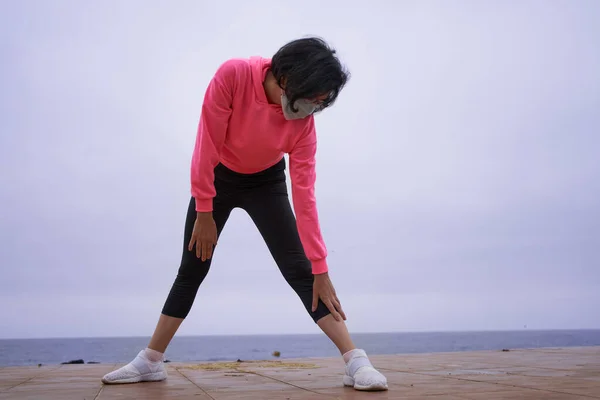 Maskeli kadın, sırtı denize dönük egzersiz yapmadan önce bacak esnetir. Yeni normal. spor oturumu