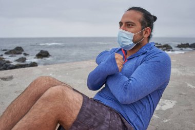 Maskeli bir adam denizin önünde mekik çeker. Yeni normallik. Spor oturumu