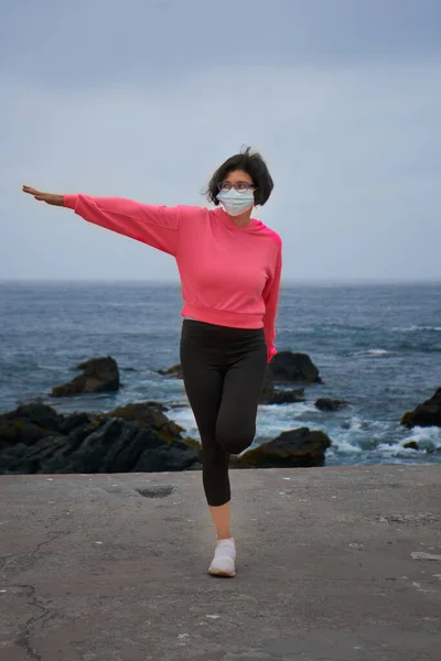 Woman with mask performs relaxation exercises in front of the sea. New normality. Sports session